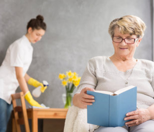 Elderly Woman Reading And Caregiver Cleaning Her Home